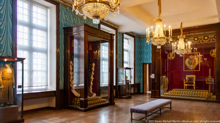 Formal museum gallery with chandeliers, blue wall hangings, glass cases, and ceremonial objects on display.