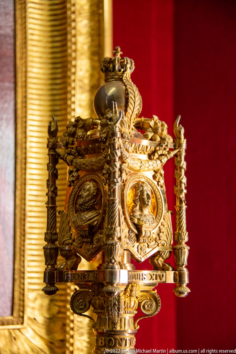 Highly ornate gilt ceremonial object with relief medallions, scrolling mounts, finials, and dark rounded top.