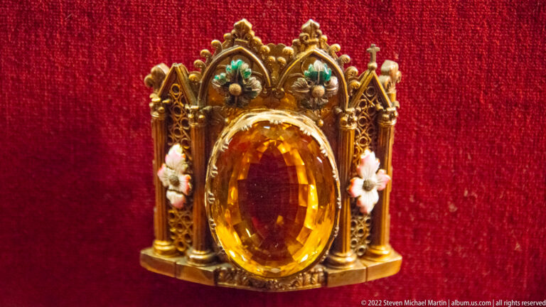 Large amber-colored oval gemstone set in a small Gothic-style gilt and enamel architectural mount.