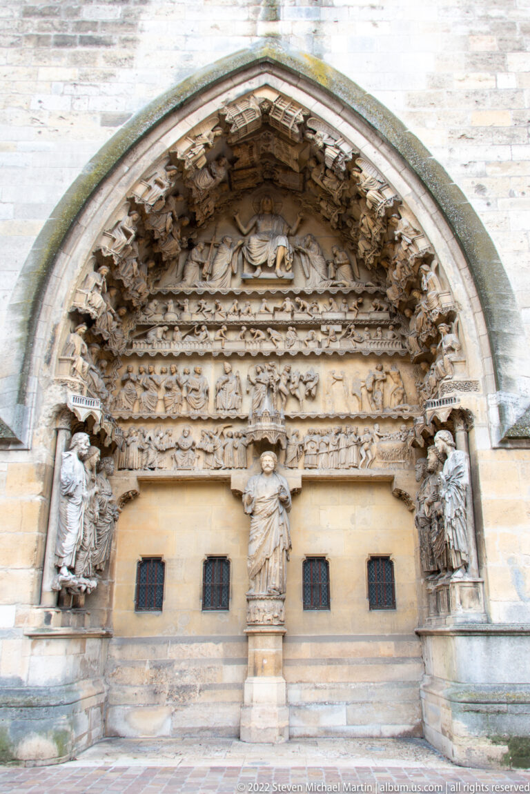 Medieval church portal with layered carved arches, sculpted tympanum, side figures, and four small rectangular openings below.