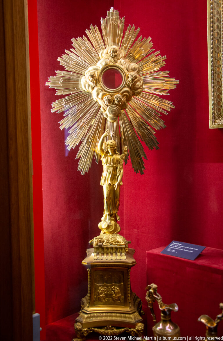 Gilt sunburst monstrance with red circular center on a sculptural stem and rectangular base.