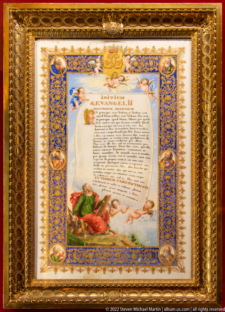 Framed illuminated document with blue-and-gold border, central text, angels, medallions, and a kneeling figure.