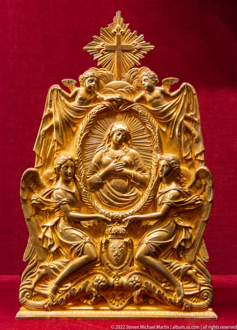 Gilt devotional relief with central bust, two seated angels, drapery, scrollwork, and a cross above.