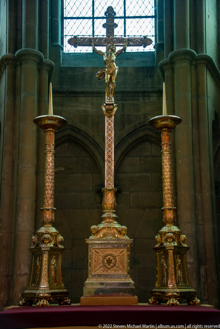 Tall crucifix centered between two matching candlesticks against a dark stone church interior.
