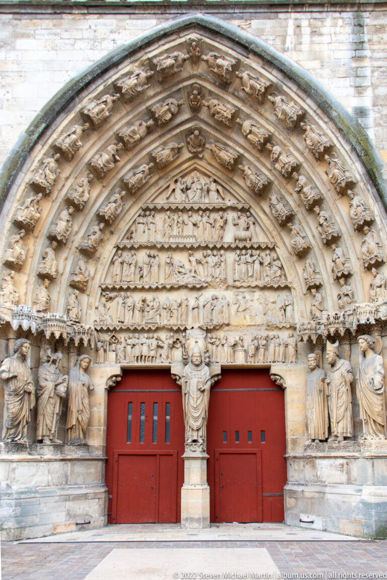 Two red church doors beneath densely carved medieval arches, tympana, and rows of standing stone figures.