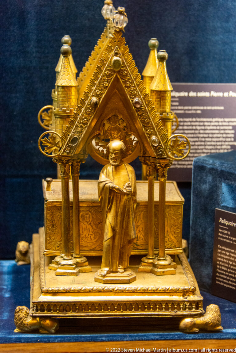 Gilt miniature shrine with gabled roof, corner turrets, and standing saint figure at the front.