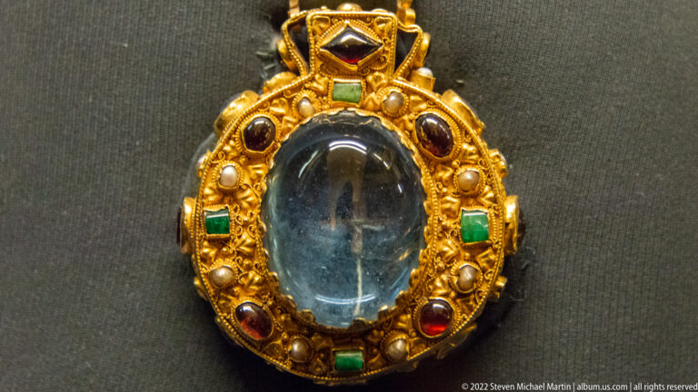 Oval transparent center in a gold mount set with pearls and red and green gemstones.