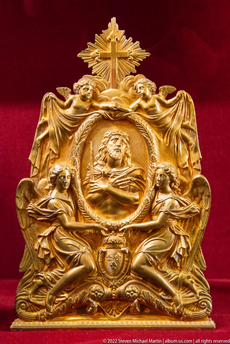 Gilt relief with central bust in a roundel, two seated angels, drapery, and radiant cross above.