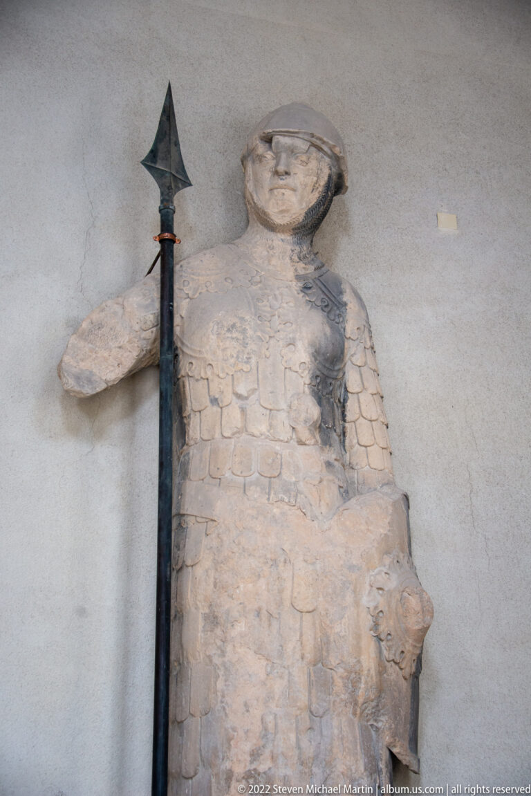 Weathered stone figure in armor standing beside a dark pole weapon with broad metal head.