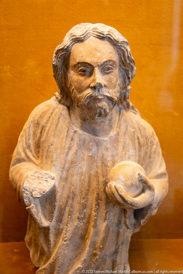 Weathered bearded stone figure holding a shallow round object against an ochre museum display background.