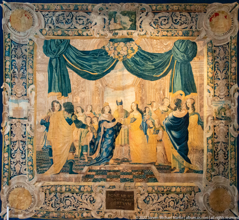 Large tapestry with a courtly gathering framed by blue drapery and a dense ornamental border.