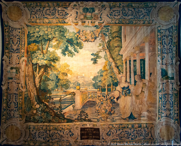 Pastoral tapestry with trees, garden landscape, bridge, classical figures, and a columned building at right.