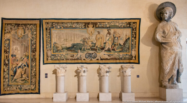 Museum wall with two tapestries, four carved stone fragments on plinths, and a standing stone figure.