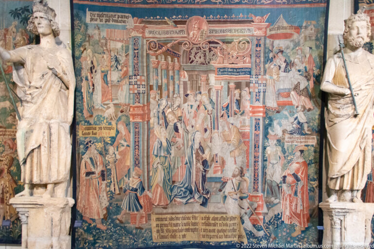 Narrative tapestry with clergy around an enthroned church figure, framed by two stone sculptures.