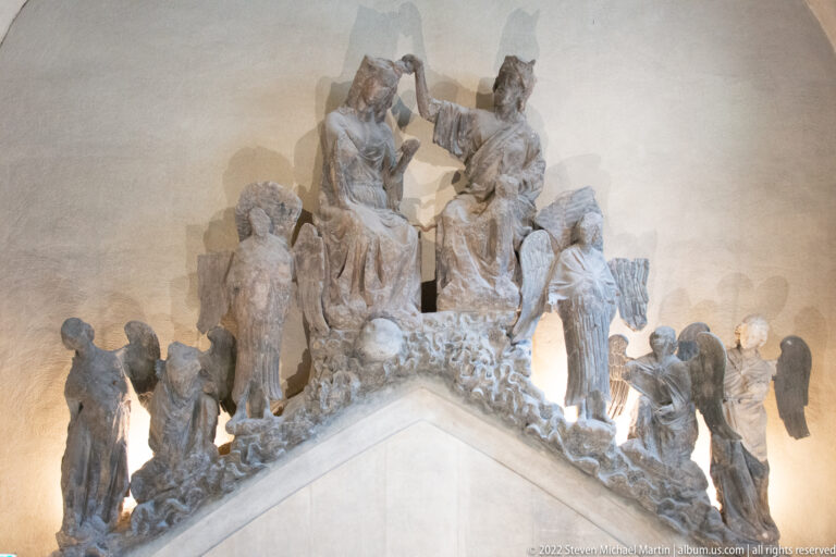 Group of weathered Gothic stone figures arranged around a peaked architectural fragment in a museum.