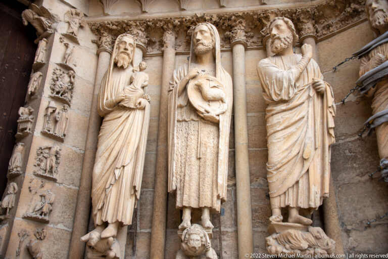 Three tall Gothic jamb statues on Reims Cathedral, with smaller narrative reliefs visible at left.