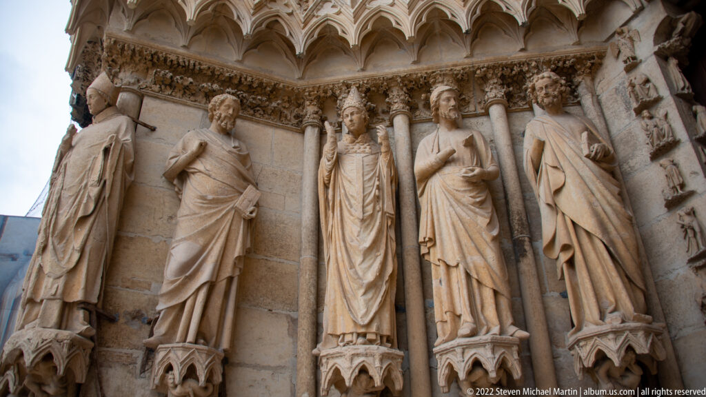 Five Gothic jamb statues in a recessed Reims Cathedral portal, including a central mitred figure.