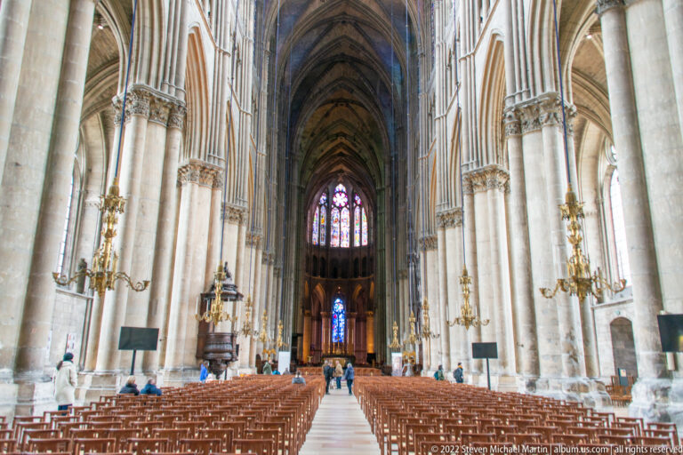 Long central nave of Reims Cathedral with rib vaults, clustered piers, rows of chairs, and stained glass beyond.