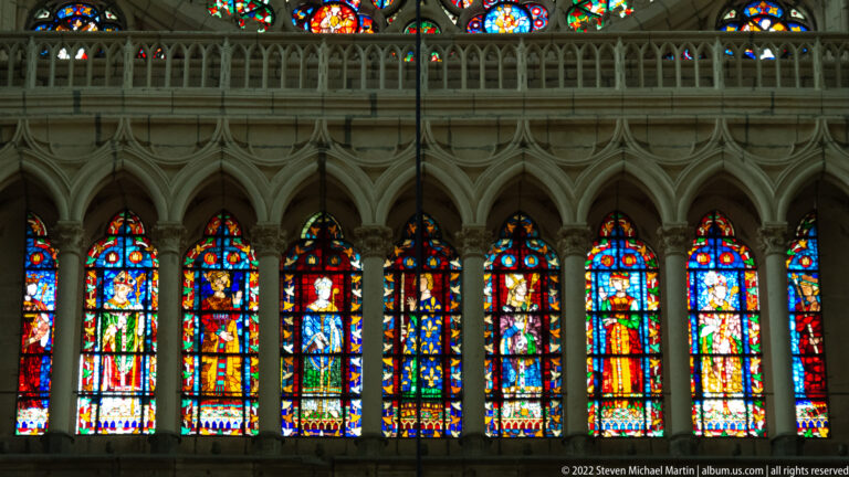 Row of tall figurative stained-glass lancets beneath a band of pointed Gothic stone arches.