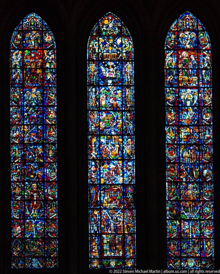Three tall stained-glass lancet windows filled with many small narrative scenes on a dark wall.
