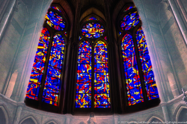 Three tall abstract stained-glass windows in blue, red, yellow, and white set within gray Gothic masonry.
