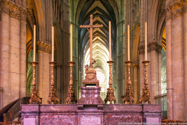 Stone altar with six tall gilded candlesticks and a suspended crucifix in a vaulted cathedral aisle.