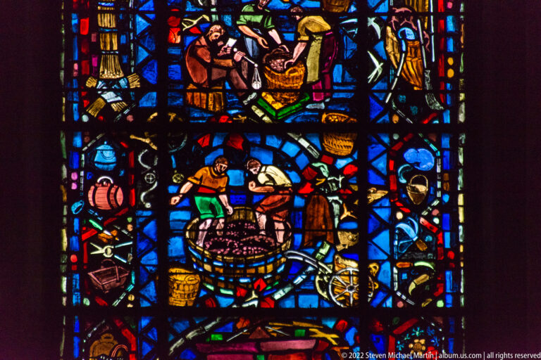 Stained-glass scene of workers processing grapes around a large vat, with baskets, barrels, and tools.