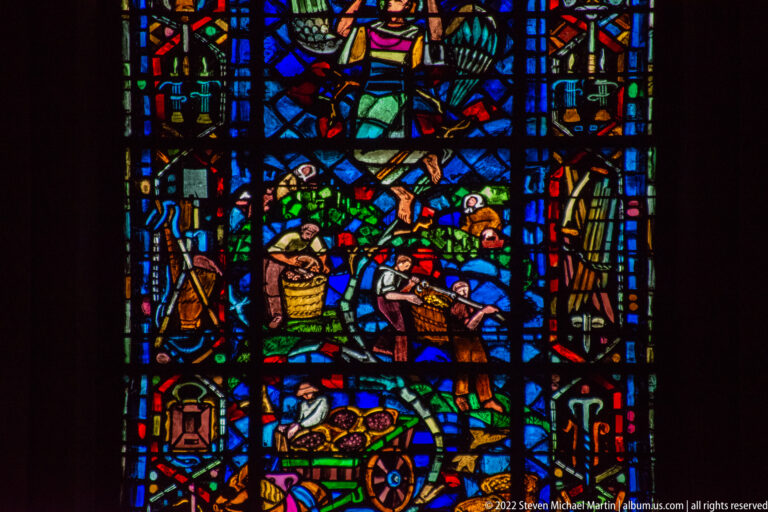 Stained-glass workers among baskets, foliage, tools, and a small cart in a blue-dominant agricultural scene.