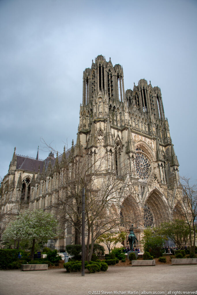 Wide view of Reims Cathedral beyond trees and an open urban foreground, with west towers and side elevation visible.