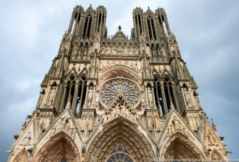 Upward view of Reims Cathedral west façade with twin towers, rose window, gables, and dense Gothic sculpture.