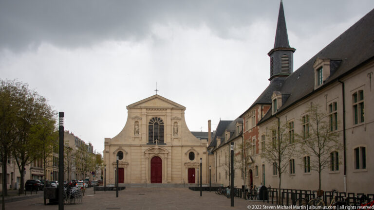 Small pale church façade with red doors beside long monastic buildings and a narrow slate spire.
