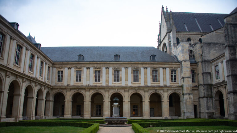 Arcaded monastic courtyard with clipped hedges, central basin, and older church masonry at right.