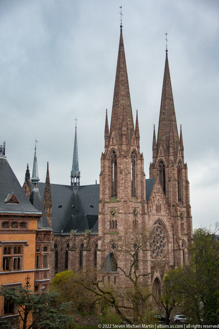 Twin-spired sandstone Gothic Revival church rises above trees and adjoining roofs under a gray sky.