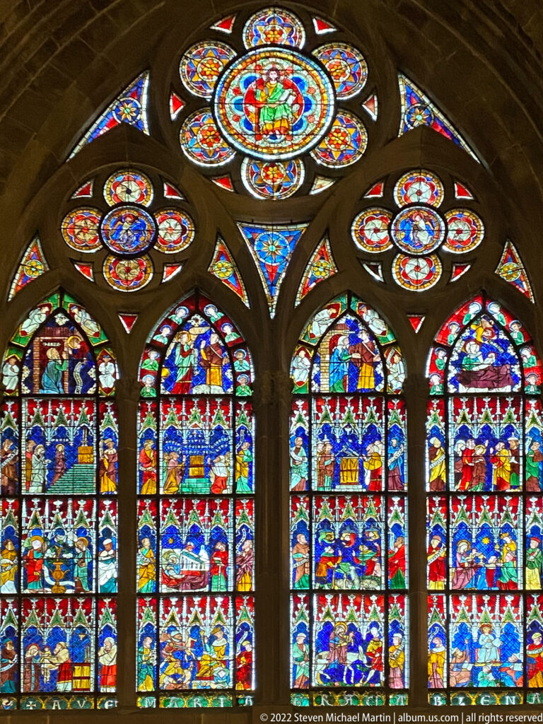 Four red-and-blue narrative stained-glass lancets beneath flower-like circular tracery windows in Strasbourg Cathedral.