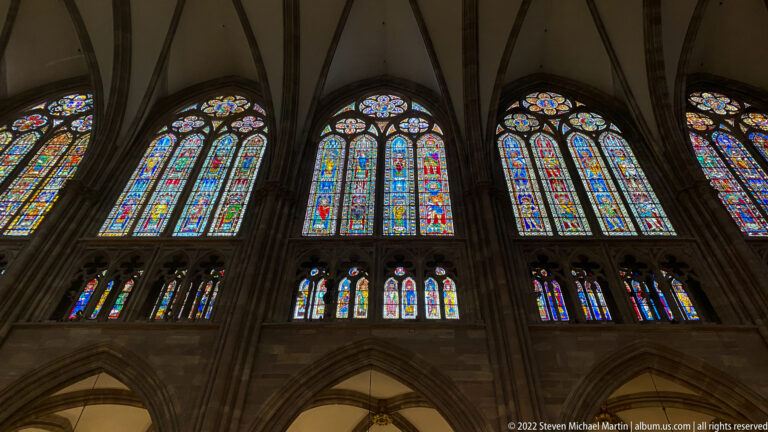 Oblique interior view of tall stained-glass windows, smaller lower lancets, ribbed vaults, and stone piers.