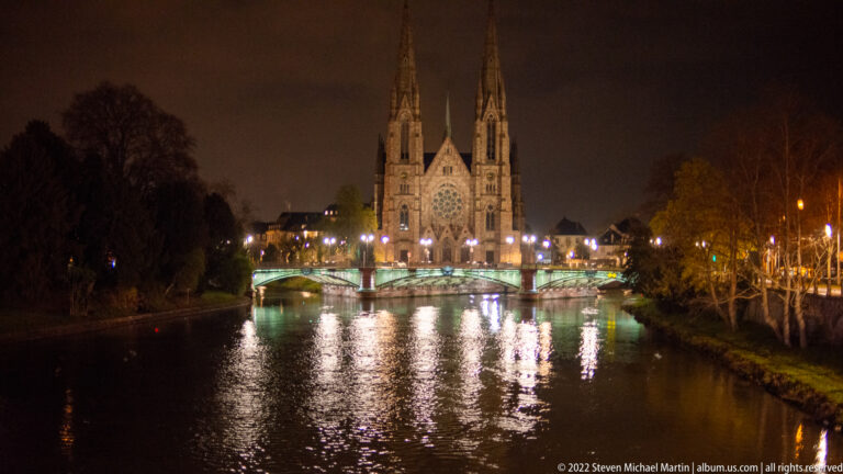 Illuminated twin-spired Saint Paul's Church beyond a bridge, reflected in the Ill River at night.