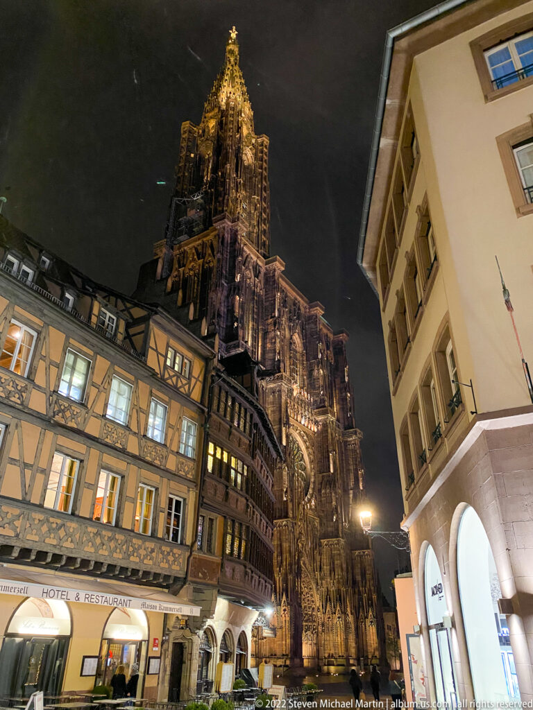 Illuminated Strasbourg Cathedral tower behind half-timbered and plastered buildings in the historic center at night.
