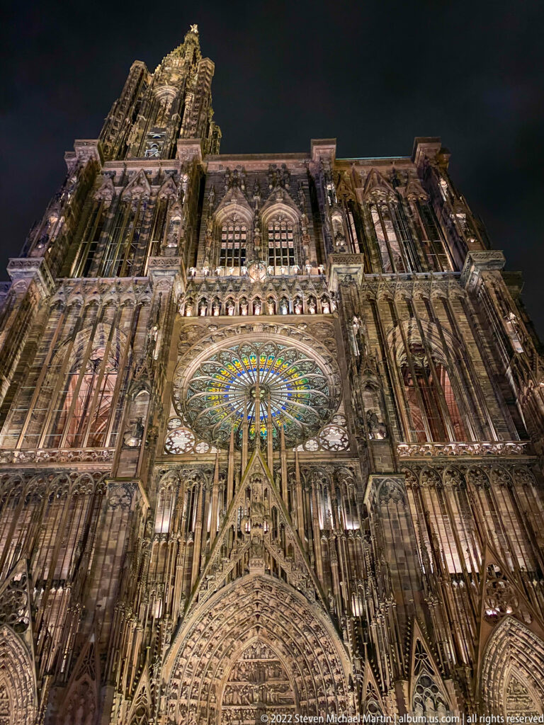 Upward nighttime view of Strasbourg Cathedral's west façade with glowing rose window, sculpted portal, and dense tracery.
