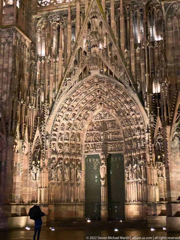 Illuminated central portal of Strasbourg Cathedral with dense Gothic sculpture, pointed gable, and a lone pedestrian for scale.
