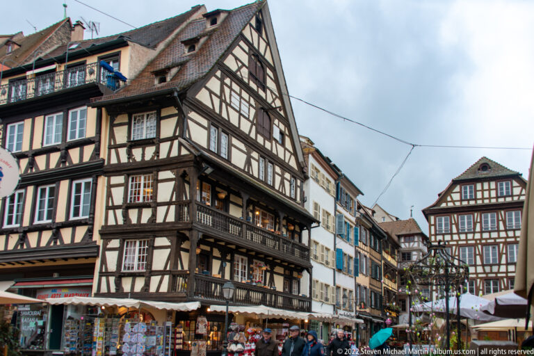 Tall half-timbered corner building overlooks a narrow Strasbourg street lined with shops and traditional façades.