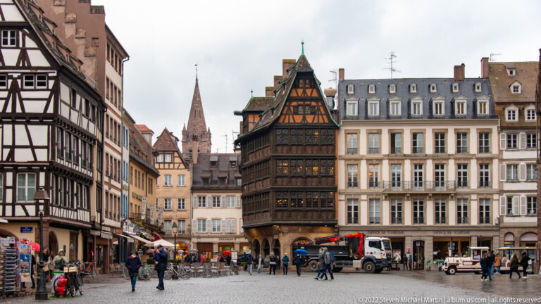 Busy Strasbourg square with an ornate dark half-timbered house and the cathedral spire visible beyond surrounding buildings.