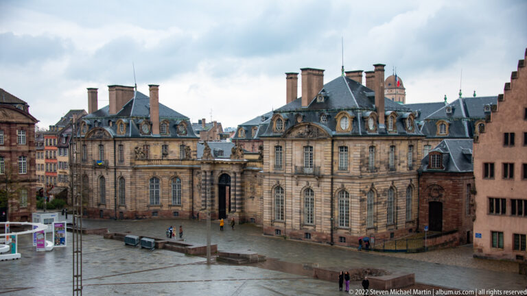 Elevated view of the Palais Rohan’s stone façades, mansard roofs, chimneys, and open courtyard with pedestrians.