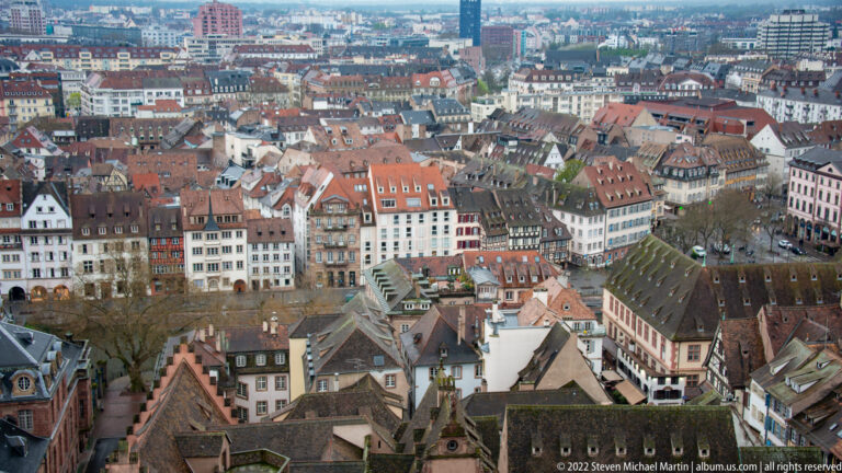 Elevated view across dense red-tiled rooftops and historic buildings in central Strasbourg, with newer districts in the distance.