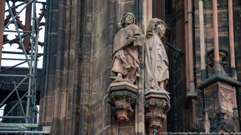 Two carved stone figures on ornate corbels beside scaffolding on the Gothic exterior of Strasbourg Cathedral.