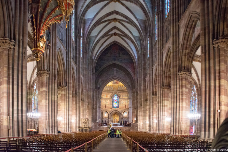 Long Gothic nave with clustered piers, ribbed vaults, stained glass, rows of chairs, and the illuminated choir beyond.