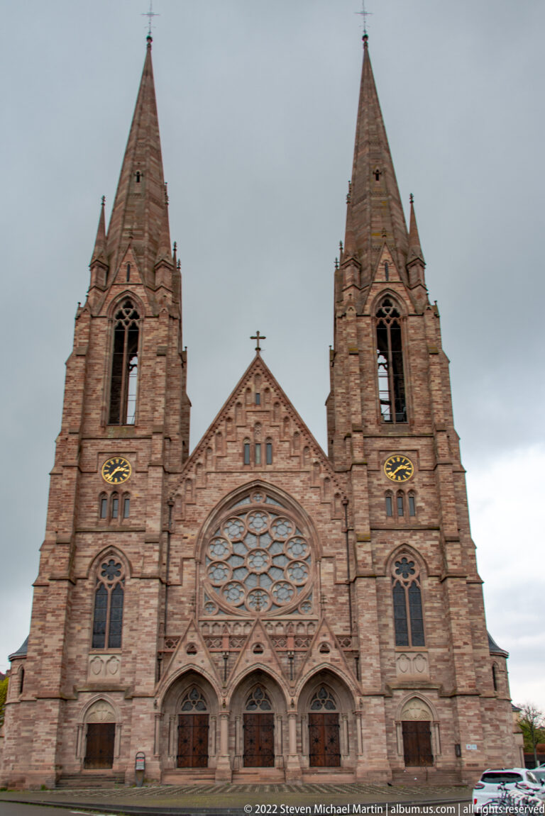 Front façade of a twin-spired sandstone Gothic Revival church with a large central rose window and pointed portals.