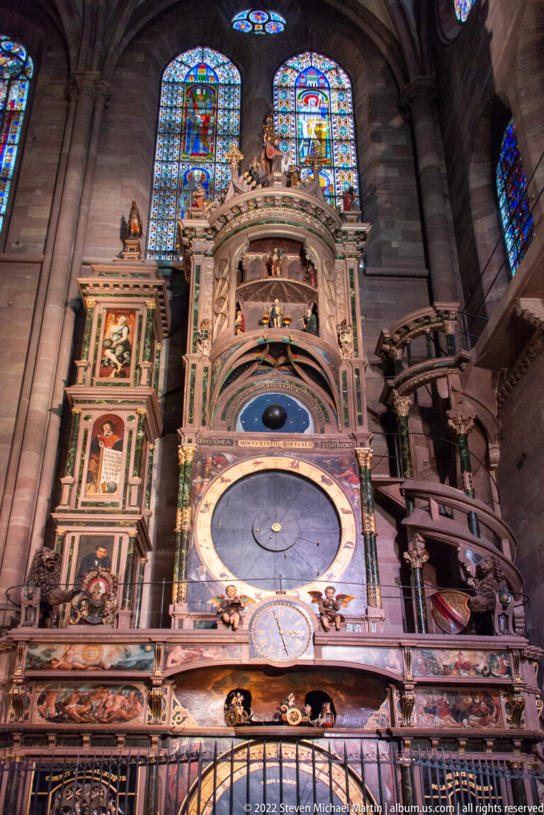 Large multilevel astronomical clock with circular dial, painted panels, sculpture, and stained glass inside Strasbourg Cathedral.