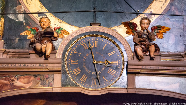 Blue Roman-numeral clock dial flanked by two winged child figures and painted architectural decoration.