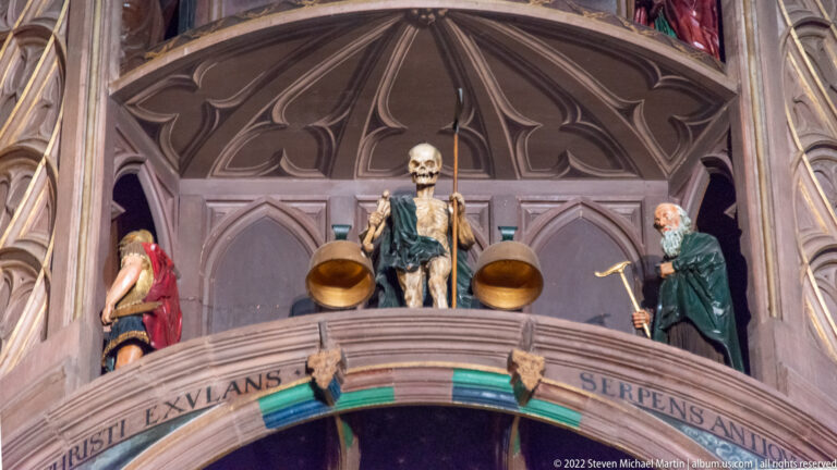Skeleton automaton stands between robed figures in a Gothic-style stage on the astronomical clock.