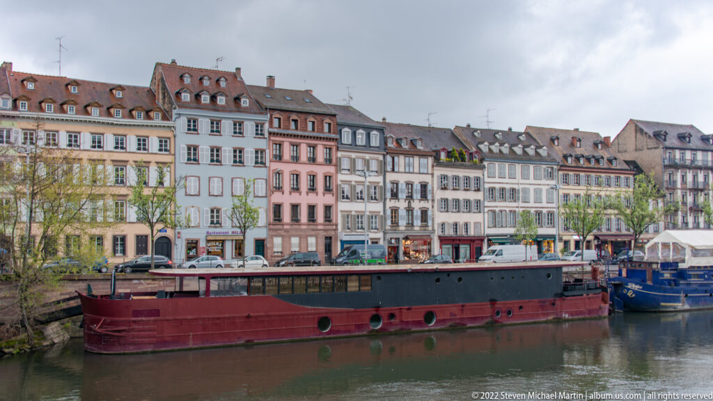 Long barge on a Strasbourg canal in front of rows of historic riverside buildings and moored boats.